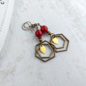 May include: Earrings with red beads, brass-colored hexagonal frames, and gold-colored leaf accents. The earrings have a vintage aesthetic and are designed to dangle. The background is a light, textured surface.