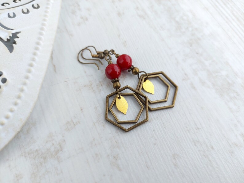 May include: Earrings with red beads, brass-colored hexagonal frames, and gold-colored leaf accents. The earrings have a vintage aesthetic and are designed to dangle. The background is a light, textured surface.