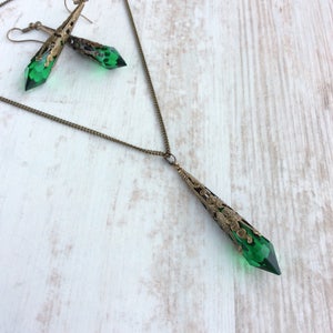 May include: A vintage-style jewelry set featuring a pendant necklace and matching earrings. The set has a bronze-colored filigree design with faceted emerald green crystal accents. The necklace and earrings are displayed on a light wood-grain surface.