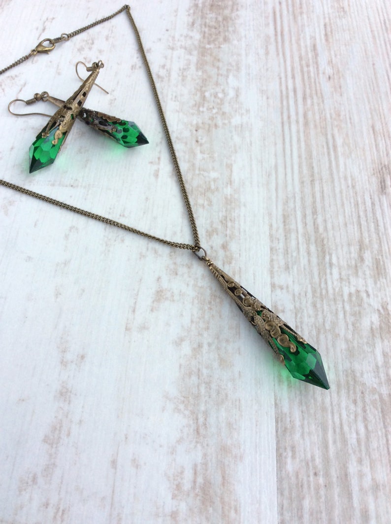 May include: A vintage-style jewelry set featuring a pendant necklace and matching earrings. The set has a bronze-colored filigree design with faceted emerald green crystal accents. The necklace and earrings are displayed on a light wood-grain surface.