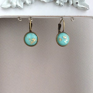 May include: A pair of round turquoise earrings with gold leaf accents. The earrings have a bronze-colored metal frame and a lever-back closure. The earrings are displayed against a neutral background.