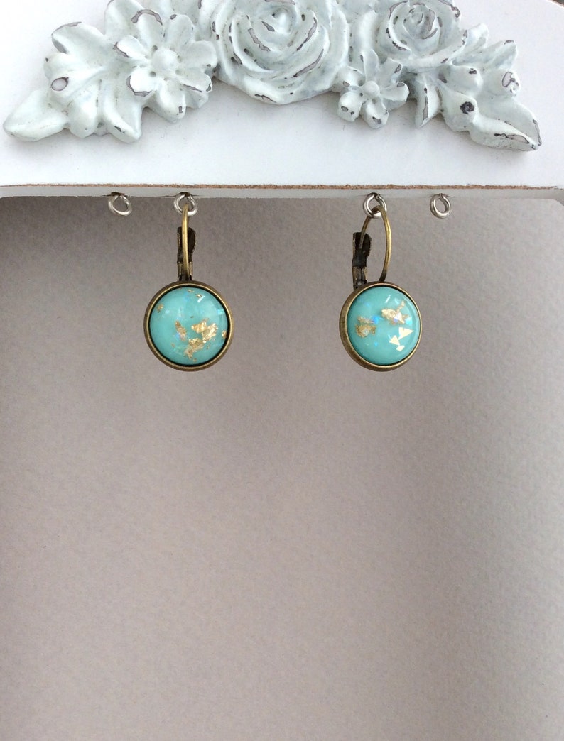 May include: A pair of round turquoise earrings with gold leaf accents. The earrings have a bronze-colored metal frame and a lever-back closure. The earrings are displayed against a neutral background.