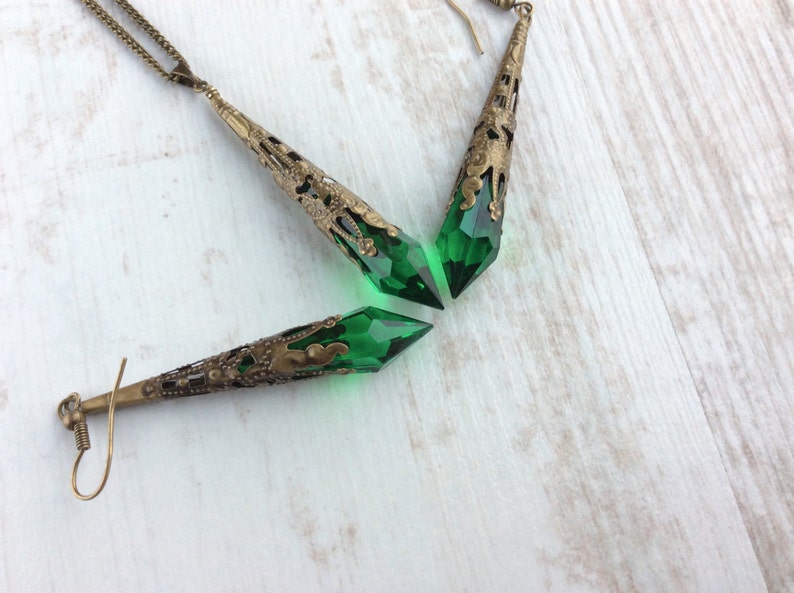 May include: A vintage-style jewelry set featuring a necklace and earrings. The set includes a long, ornate pendant and two earrings with a bronze-colored metal filigree design. Each piece holds a faceted, emerald-green crystal, creating a striking contrast.