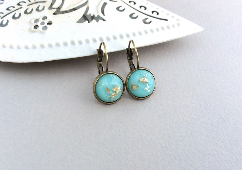 May include: A pair of round turquoise earrings with gold leaf accents. The earrings have a bronze-colored metal frame and a lever-back closure. The earrings are displayed on a white decorative surface.