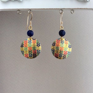 May include: Gold-toned dangle earrings with a circular design. Each earring features a navy blue bead and a colorful, leaf-patterned disc. The leaves are in shades of red, orange, green, and blue on a gold background.