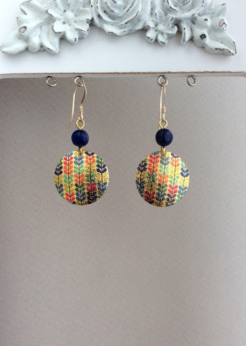 May include: Gold-toned dangle earrings with a circular design. Each earring features a navy blue bead and a colorful, leaf-patterned disc. The leaves are in shades of red, orange, green, and blue on a gold background.