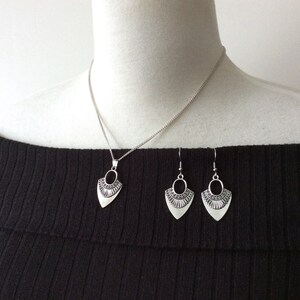 May include: Silver-toned jewelry set featuring a pendant necklace and matching earrings. The pendant and earrings have a stylized, shield-like shape with an oval cutout and decorative detailing. The necklace has a delicate chain.