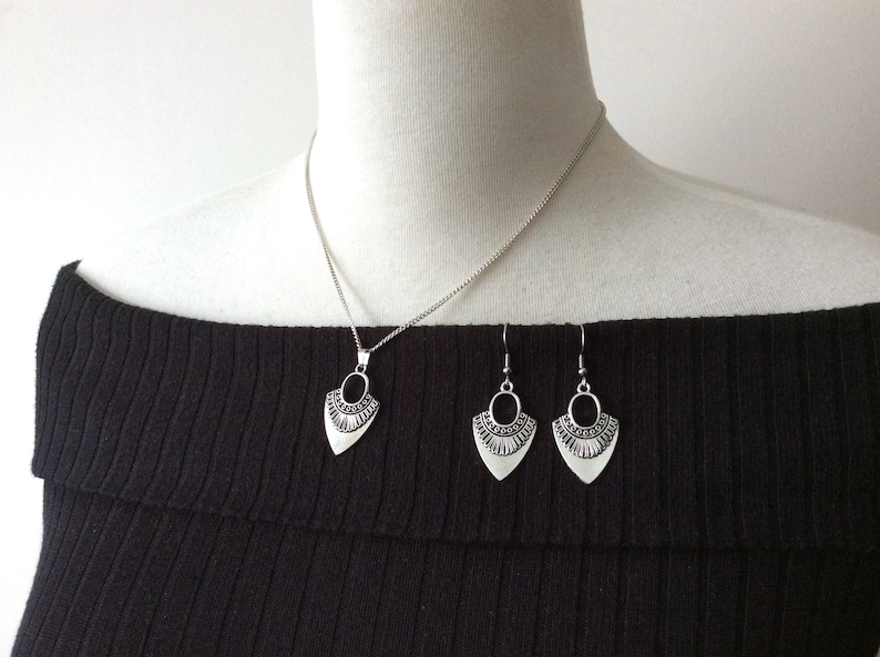 May include: Silver-toned jewelry set featuring a pendant necklace and matching earrings. The pendant and earrings have a stylized, shield-like shape with an oval cutout and decorative detailing. The necklace has a delicate chain.