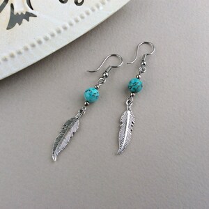 May include: A pair of dangle earrings featuring turquoise beads and silver-colored feather charms. The earrings have a hook closure and are displayed on a light gray surface. The earrings are a jewelry item.