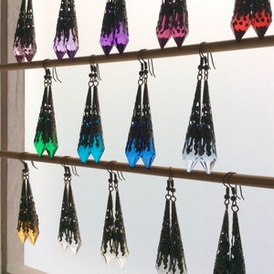 May include: A collection of dangle earrings in various colors, including pink, purple, green, blue, and clear. Each earring features a filigree metalwork design with a teardrop-shaped, faceted crystal. The earrings are displayed on wooden rods.