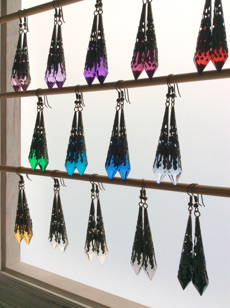 May include: A collection of dangle earrings in various colors, including pink, purple, green, blue, and clear. Each earring features a filigree metalwork design with a teardrop-shaped, faceted crystal. The earrings are displayed on wooden rods.
