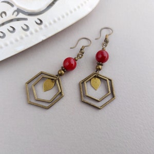 May include: A pair of dangle earrings with a geometric design. Each earring features a red bead, brass-colored metal accents, and a double hexagon shape with a leaf charm. The earrings are displayed on a white surface.