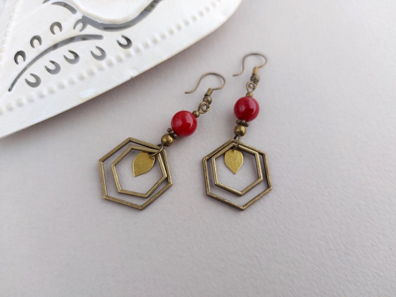 May include: A pair of dangle earrings with a geometric design. Each earring features a red bead, brass-colored metal accents, and a double hexagon shape with a leaf charm. The earrings are displayed on a white surface.