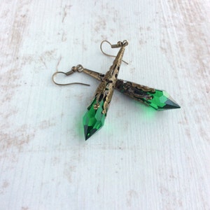 May include: A pair of dangle earrings featuring faceted green crystal pendants. The earrings have an antique brass filigree design at the top and a hook closure. The earrings are displayed on a white wooden surface.