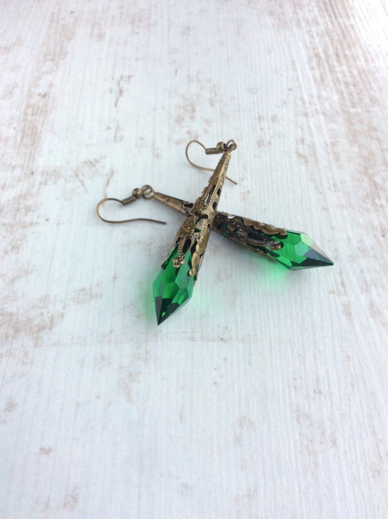 May include: A pair of dangle earrings featuring faceted green crystal pendants. The earrings have an antique brass filigree design at the top and a hook closure. The earrings are displayed on a white wooden surface.