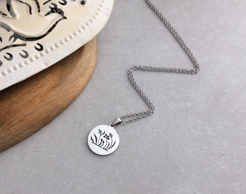 Puede incluir: Un collar plateado con un colgante redondo con un dise&ntilde;o floral negro. El colgante cuelga de una cadena delicada. El collar se muestra sobre una superficie gris.