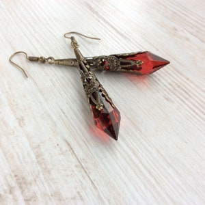May include: A pair of long, ornate earrings with faceted, deep red crystal pendants. The earrings have an antique brass finish with intricate filigree detailing. The crystals are pointed at the bottom and are approximately 2 inches long.