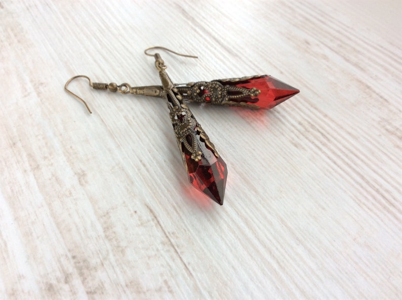 May include: A pair of long, ornate earrings with faceted, deep red crystal pendants. The earrings have an antique brass finish with intricate filigree detailing. The crystals are pointed at the bottom and are approximately 2 inches long.