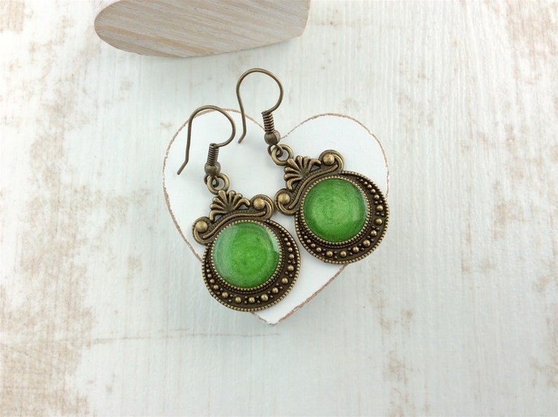 May include: A pair of antique-style earrings with a green center and bronze-colored filigree. The earrings have a circular design with a beaded border and decorative flourishes. The earrings are displayed on a white heart-shaped object.