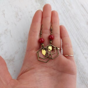 May include: A pair of handmade earrings featuring red beads, brass-colored hexagonal frames, and small leaf-shaped accents. The earrings have a vintage aesthetic and are designed to dangle. The jewelry is displayed against a light-colored background.