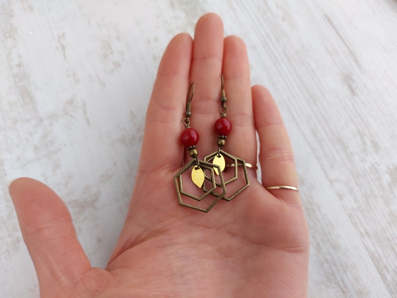 May include: A pair of handmade earrings featuring red beads, brass-colored hexagonal frames, and small leaf-shaped accents. The earrings have a vintage aesthetic and are designed to dangle. The jewelry is displayed against a light-colored background.