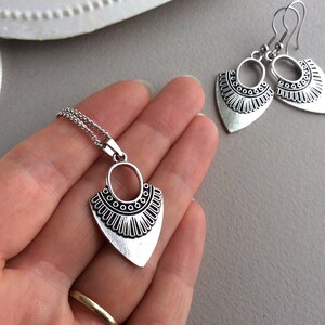 May include: A silver-toned jewelry set featuring a pendant necklace and matching earrings. The pendant and earrings have a stylized, geometric design with an oval top and a triangular bottom, adorned with decorative detailing. The set is displayed on a neutral background.