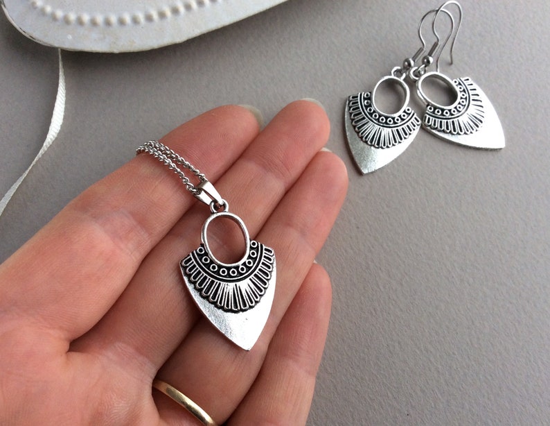 May include: A silver-toned jewelry set featuring a pendant necklace and matching earrings. The pendant and earrings have a stylized, geometric design with an oval top and a triangular bottom, adorned with decorative detailing. The set is displayed on a neutral background.