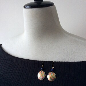 May include: A pair of round earrings with a colorful, patterned design. The earrings have a gold-colored metal backing and a small, dark blue bead. The earrings are hanging on a white mannequin.