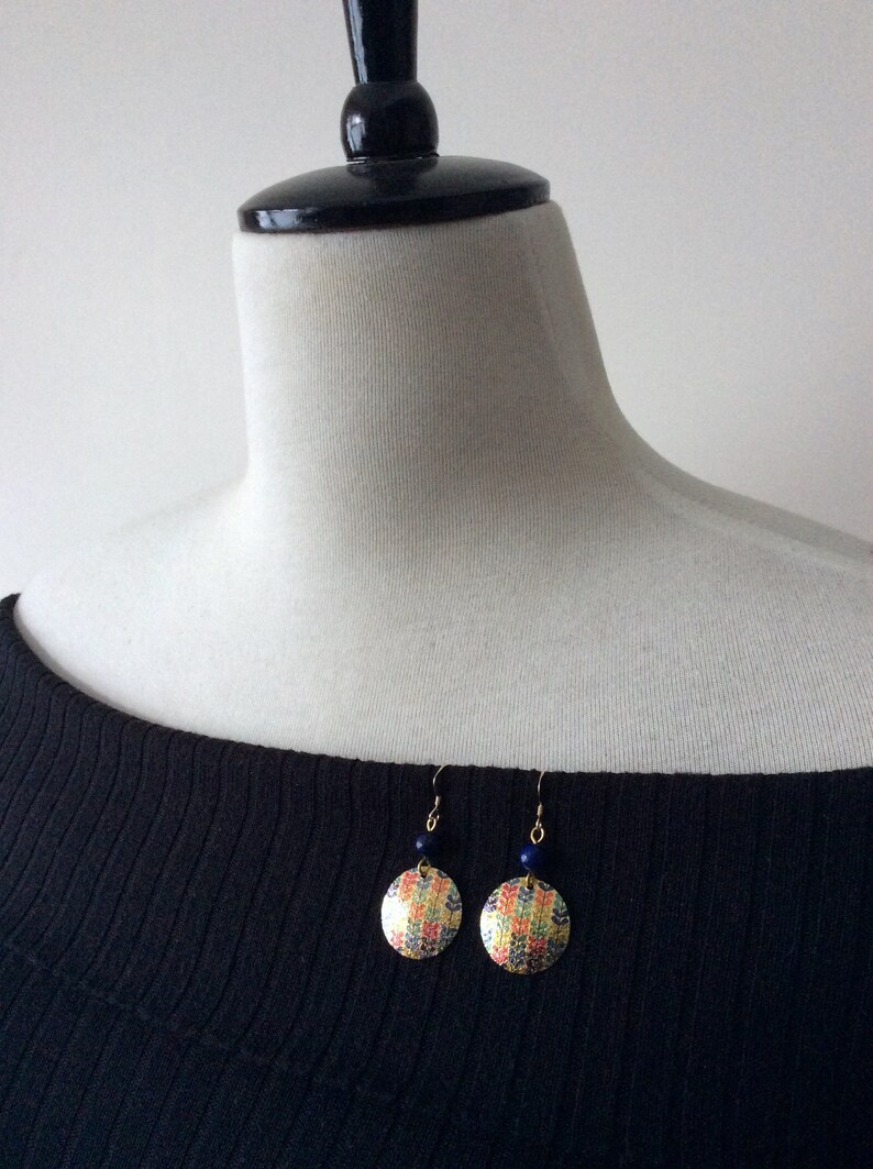 May include: A pair of round earrings with a colorful, patterned design. The earrings have a gold-colored metal backing and a small, dark blue bead. The earrings are hanging on a white mannequin.