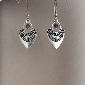 May include: A pair of silver-toned earrings with a tribal design. Each earring features a teardrop-shaped pendant with a decorative border and a small purple bead. The earrings hang from a simple hook.