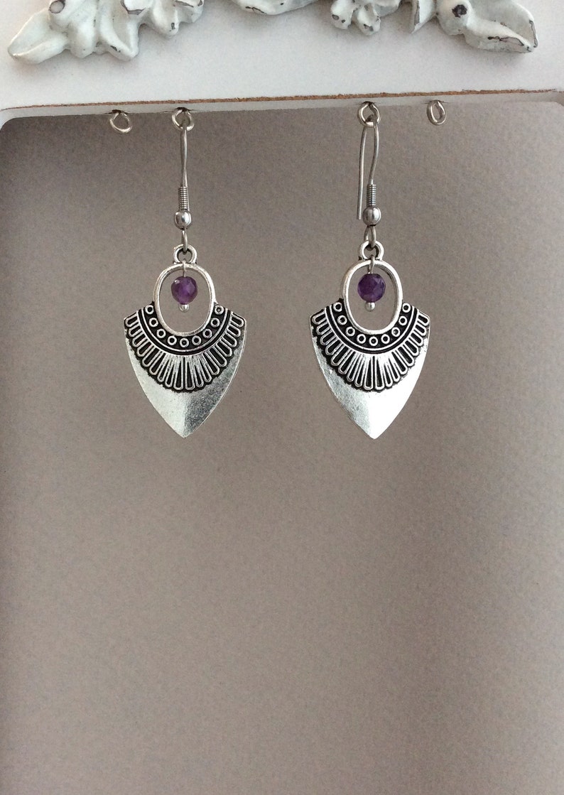 May include: A pair of silver-toned earrings with a tribal design. Each earring features a teardrop-shaped pendant with a decorative border and a small purple bead. The earrings hang from a simple hook.