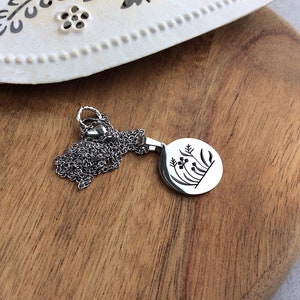 Puede incluir: Un collar con colgante plateado con un colgante redondo con un dise&ntilde;o floral. El colgante cuelga de una cadena delicada. El collar se muestra sobre una superficie de madera.