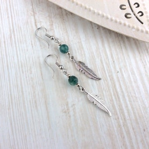 May include: A pair of dangle earrings featuring a silver feather charm and a green, round bead. The earrings have a silver hook closure. The jewelry is displayed on a light wood surface.