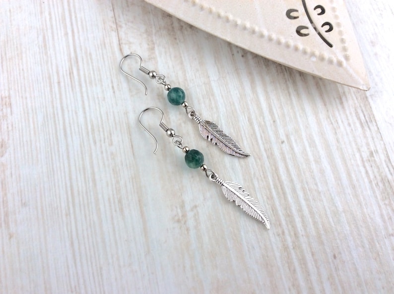 May include: A pair of dangle earrings featuring a silver feather charm and a green, round bead. The earrings have a silver hook closure. The jewelry is displayed on a light wood surface.