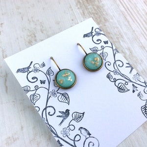 May include: A pair of round turquoise earrings with gold leaf accents, set in a bronze-colored metal. The earrings are displayed on a white card with a black floral and bird design. The background is a light wood surface.