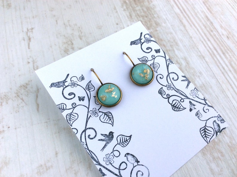 May include: A pair of round turquoise earrings with gold leaf accents, set in a bronze-colored metal. The earrings are displayed on a white card with a black floral and bird design. The background is a light wood surface.