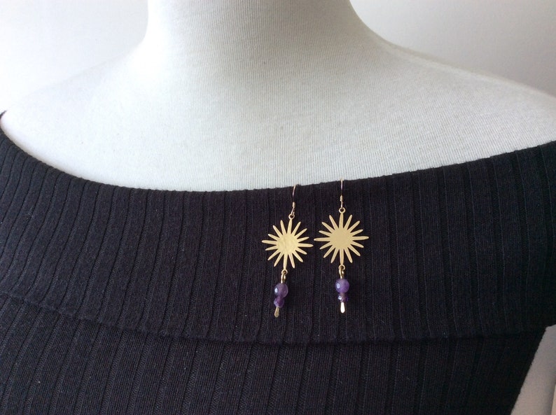 May include: Gold-colored sunburst earrings with a dangling cluster of purple beads. The earrings are suspended from a simple hook and are displayed on a white surface against a black ribbed fabric.