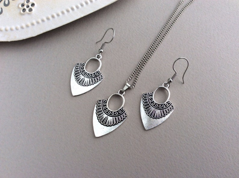 May include: A silver-toned jewelry set featuring a necklace and matching earrings. The set includes a pendant necklace and two dangle earrings, all with a decorative, geometric design. The jewelry is displayed on a neutral gray surface.