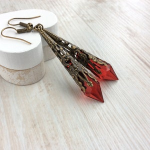May include: A pair of long, ornate earrings with a vintage aesthetic. The earrings feature a bronze-colored filigree design and are accented with faceted, red-colored crystal pendants. The earrings are displayed on a white surface.