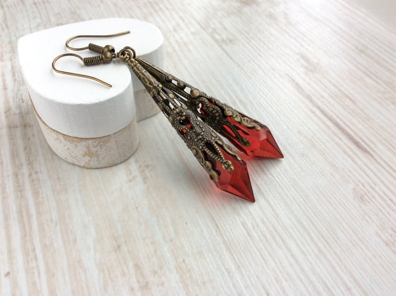 May include: A pair of long, ornate earrings with a vintage aesthetic. The earrings feature a bronze-colored filigree design and are accented with faceted, red-colored crystal pendants. The earrings are displayed on a white surface.