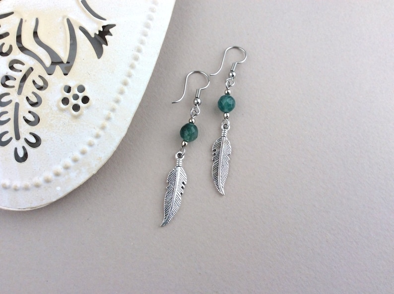 May include: A pair of dangle earrings featuring silver-colored feather charms and green gemstone beads. The earrings have a hook closure and are displayed against a neutral background. The earrings are a bohemian style.