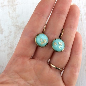 May include: A pair of round, turquoise earrings with gold leaf accents. The earrings have a bronze-colored metal frame and a lever-back closure. The earrings are approximately 0.75 inches in diameter.