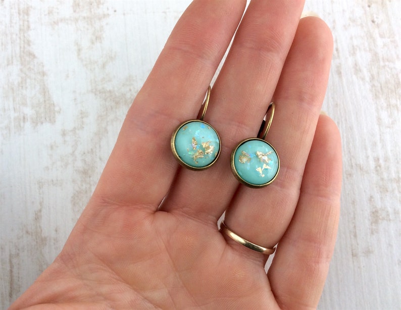 May include: A pair of round, turquoise earrings with gold leaf accents. The earrings have a bronze-colored metal frame and a lever-back closure. The earrings are approximately 0.75 inches in diameter.