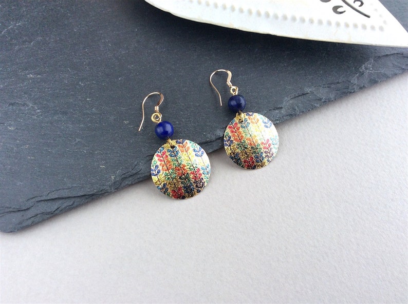 May include: A pair of gold-toned dangle earrings. Each earring features a round, patterned pendant with gold, red, blue, and yellow hues, paired with a small, dark blue bead. The earrings hang from gold-colored hooks.