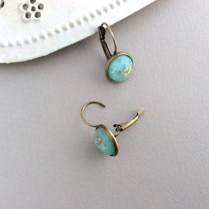 May include: A pair of turquoise leverback earrings with gold flecks. The earrings have a round, bezel-set design and a bronze-colored metal finish. The earrings are displayed on a light gray surface.