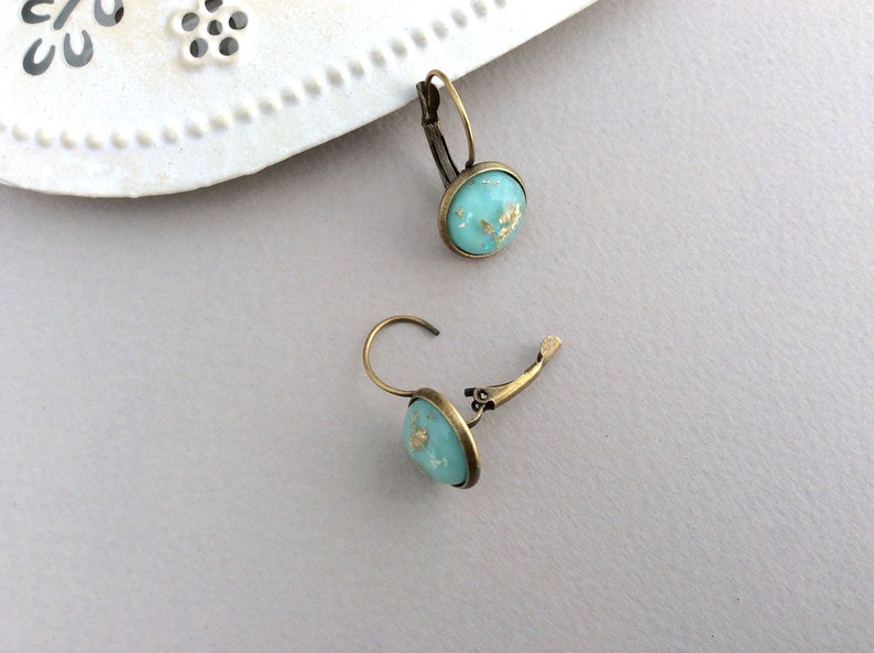May include: A pair of turquoise leverback earrings with gold flecks. The earrings have a round, bezel-set design and a bronze-colored metal finish. The earrings are displayed on a light gray surface.
