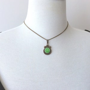 May include: A vintage-style pendant necklace featuring a round, light green stone set in an ornate, bronze-toned metal frame. The pendant hangs from a delicate chain, creating a classic and elegant look. The necklace is displayed on a white mannequin.