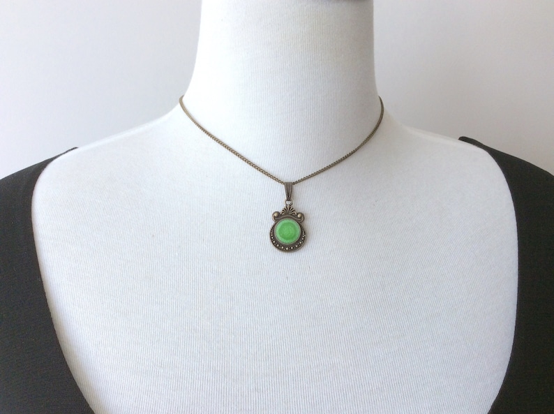 May include: A vintage-style pendant necklace featuring a round, light green stone set in an ornate, bronze-toned metal frame. The pendant hangs from a delicate chain, creating a classic and elegant look. The necklace is displayed on a white mannequin.