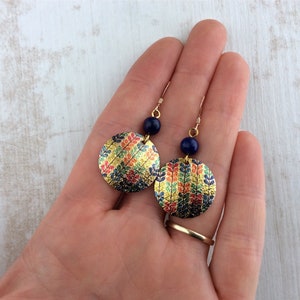May include: Gold-toned dangle earrings with a circular design. The earrings feature a colorful floral pattern and a dark blue bead. The earrings are held in a hand.