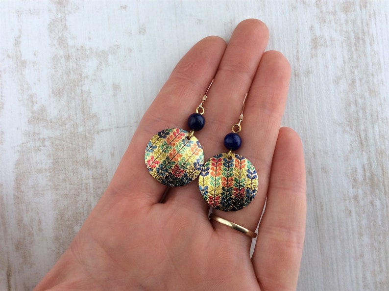 May include: Gold-toned dangle earrings with a circular design. The earrings feature a colorful floral pattern and a dark blue bead. The earrings are held in a hand.